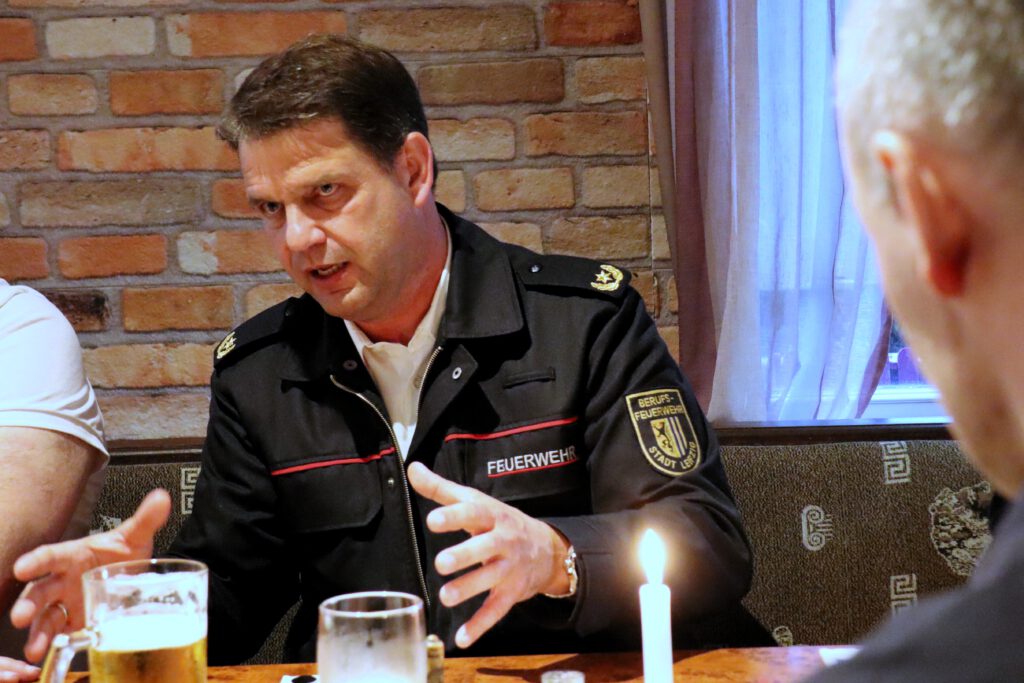 Branddirektor Axel Schuh in der Frontale. Er trägt seine Feuerwehruniform mit dem Leipiger Stadtwappen am Oberarm und einem goldenen Stern auf den Schulterklappen. Er gestikuliert, während er den Tischnachbarn erzählt.